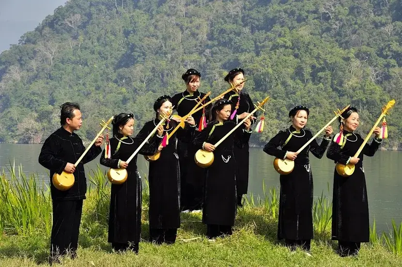 A group of Tay ethnic people in traditional black indigo tunics playing the Dan Tinh (gourd lute) by a lake in Ha Giang, Vietnam.