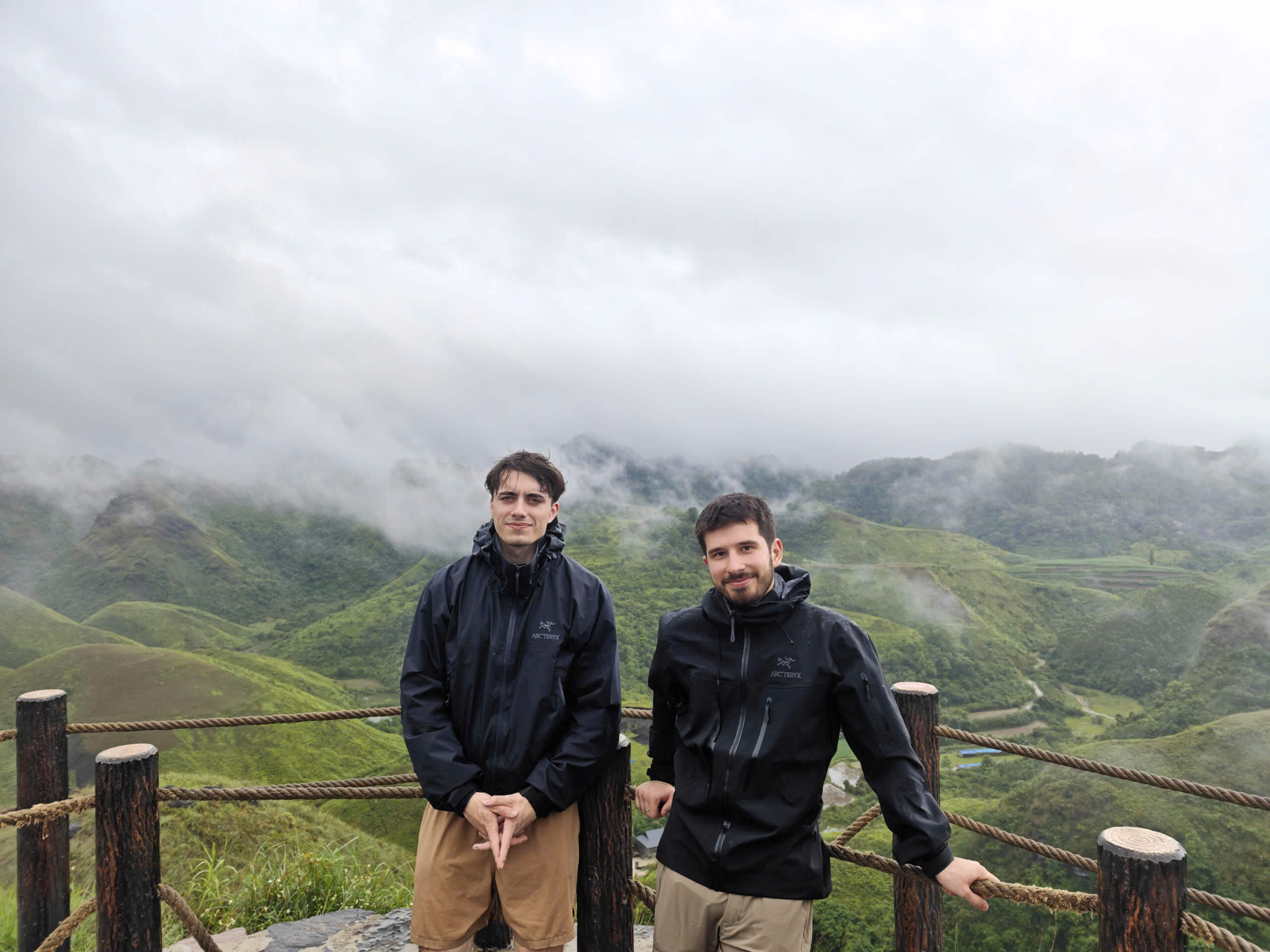 Two male travelers wearing light jackets standing at a scenic wooden viewpoint, surrounded by lush green hills and low-hanging misty clouds during a mountain ride in Northern Vietnam.