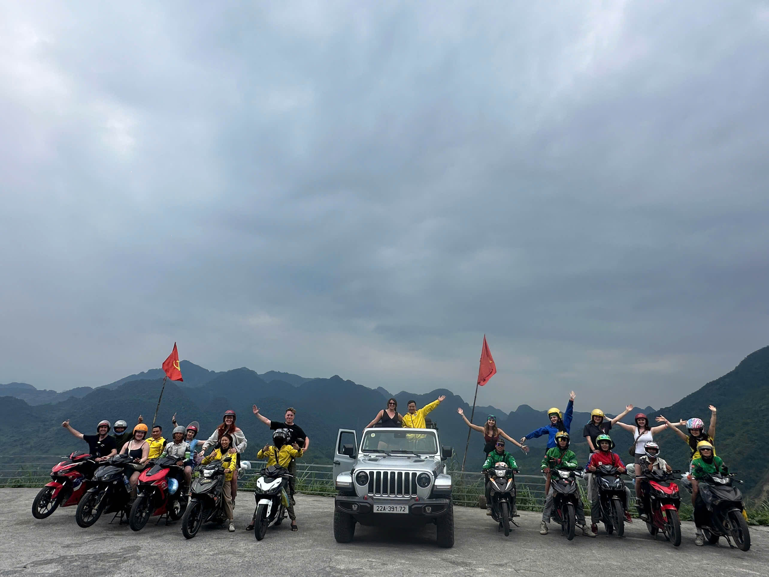 A large group of excited Motorventures travelers, Easy Riders on motorbikes, and an open-top Jeep posing together at a scenic mountain viewpoint on the Northern Vietnam loop.