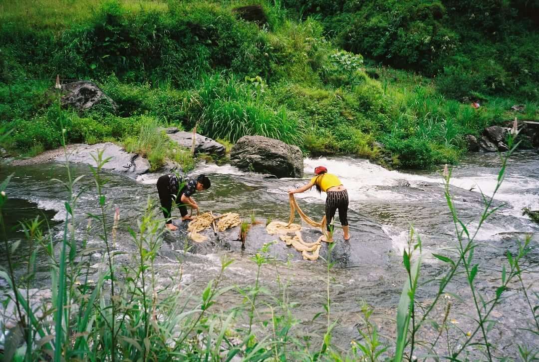 Two local ethnic women washing traditional hemp fabric in a clear, rocky stream surrounded by lush green nature during a cultural stop on the Ha Giang loop.