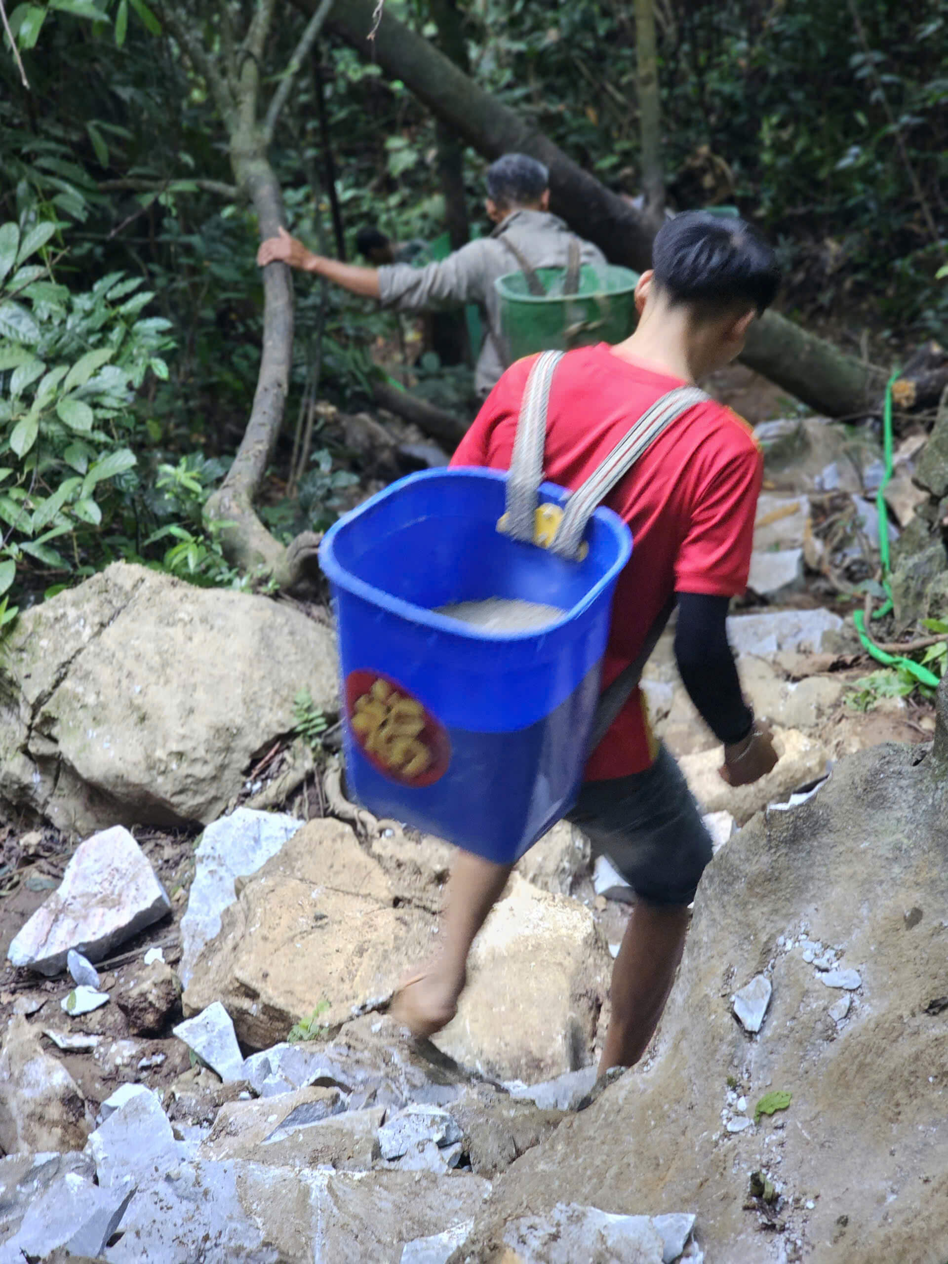 A local worker carrying a large green basket on his back, carefully navigating down the old, steep, and rocky jungle trail during the early stages of the Khau Lan Waterfall path construction.