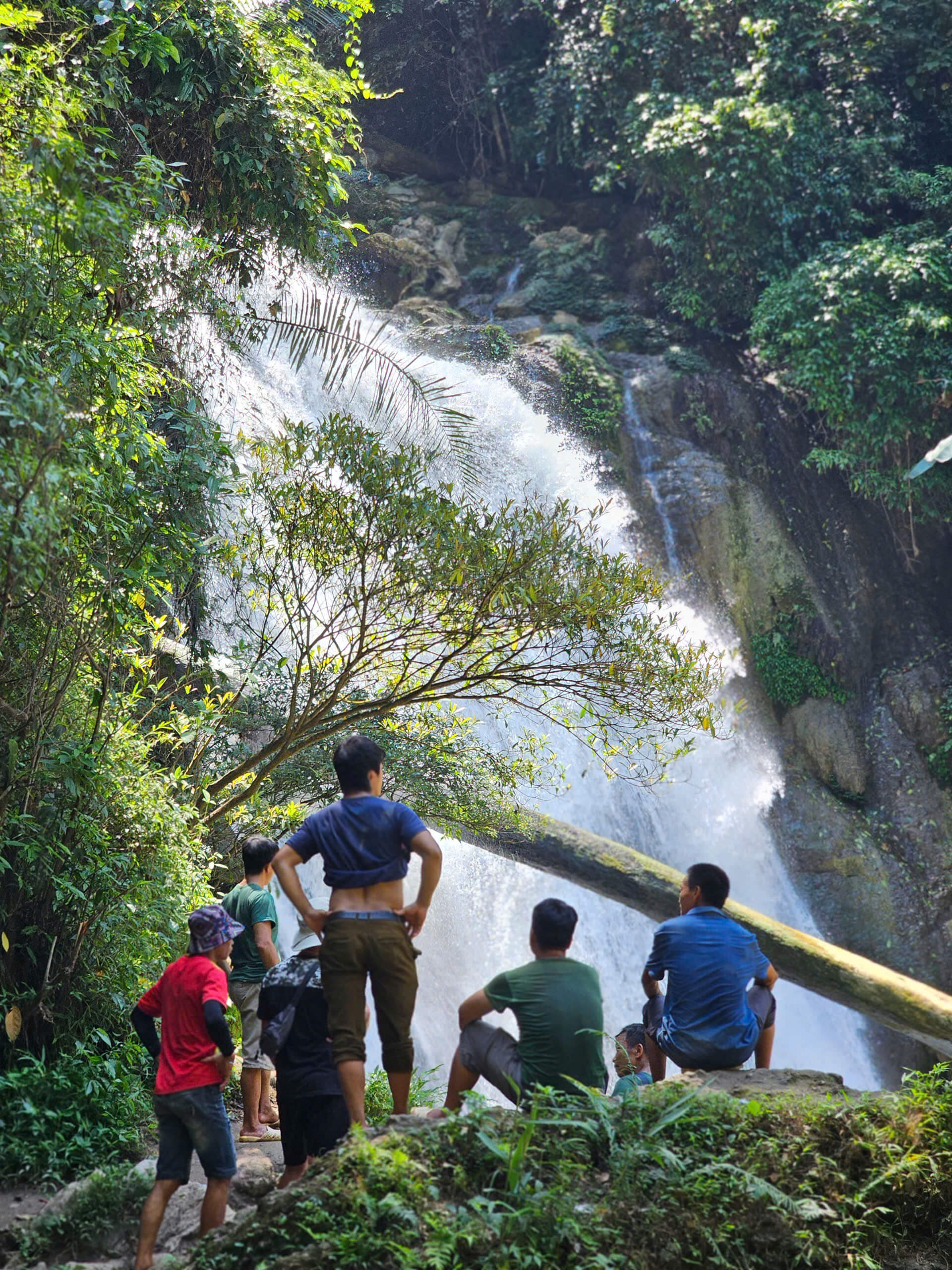 A group of local men standing on a dirt edge in the lush green jungle, admiring the beautiful, cascading waters of Khau Lan Waterfall in Ha Giang.