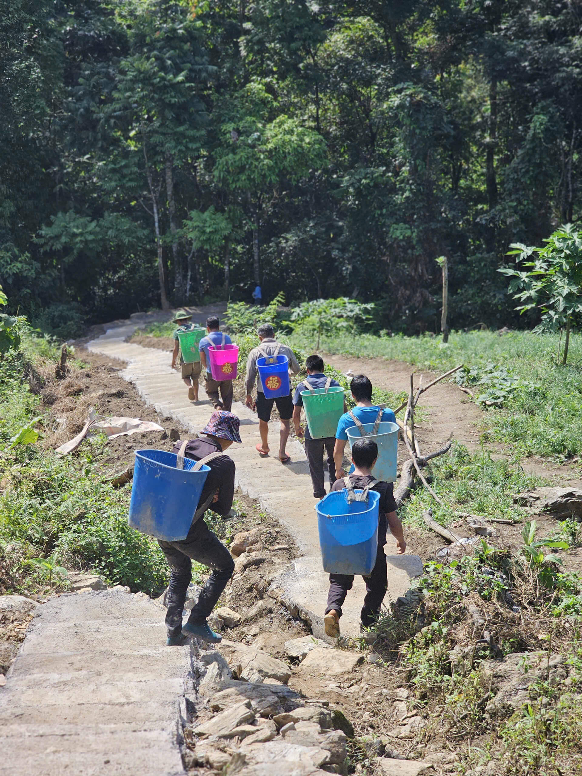 A line of local men carrying heavy blue and green buckets on their backs, walking down the newly poured, smooth concrete path leading to Khau Lan Waterfall in Ha Giang.