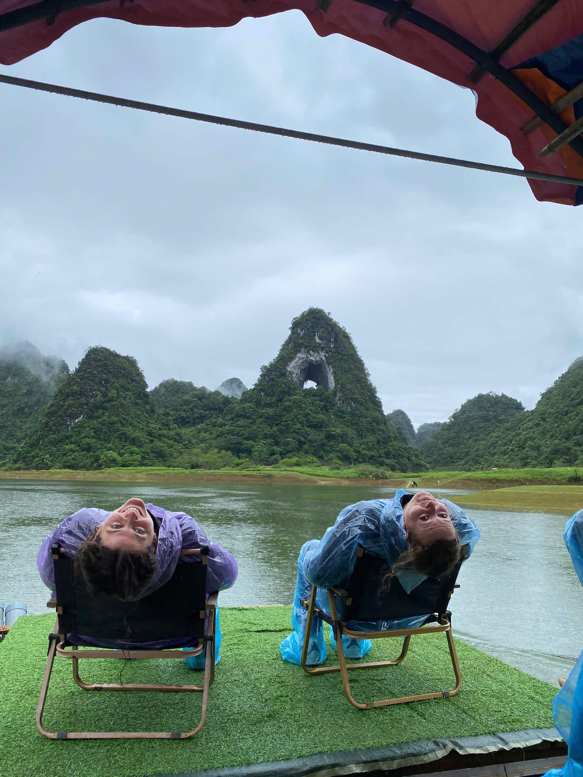 Two female tourists wearing blue and purple raincoats leaning back playfully on camping chairs on a raft, with the famous Angel Eye Mountain (Nui Thung) in the background under a cloudy sky.