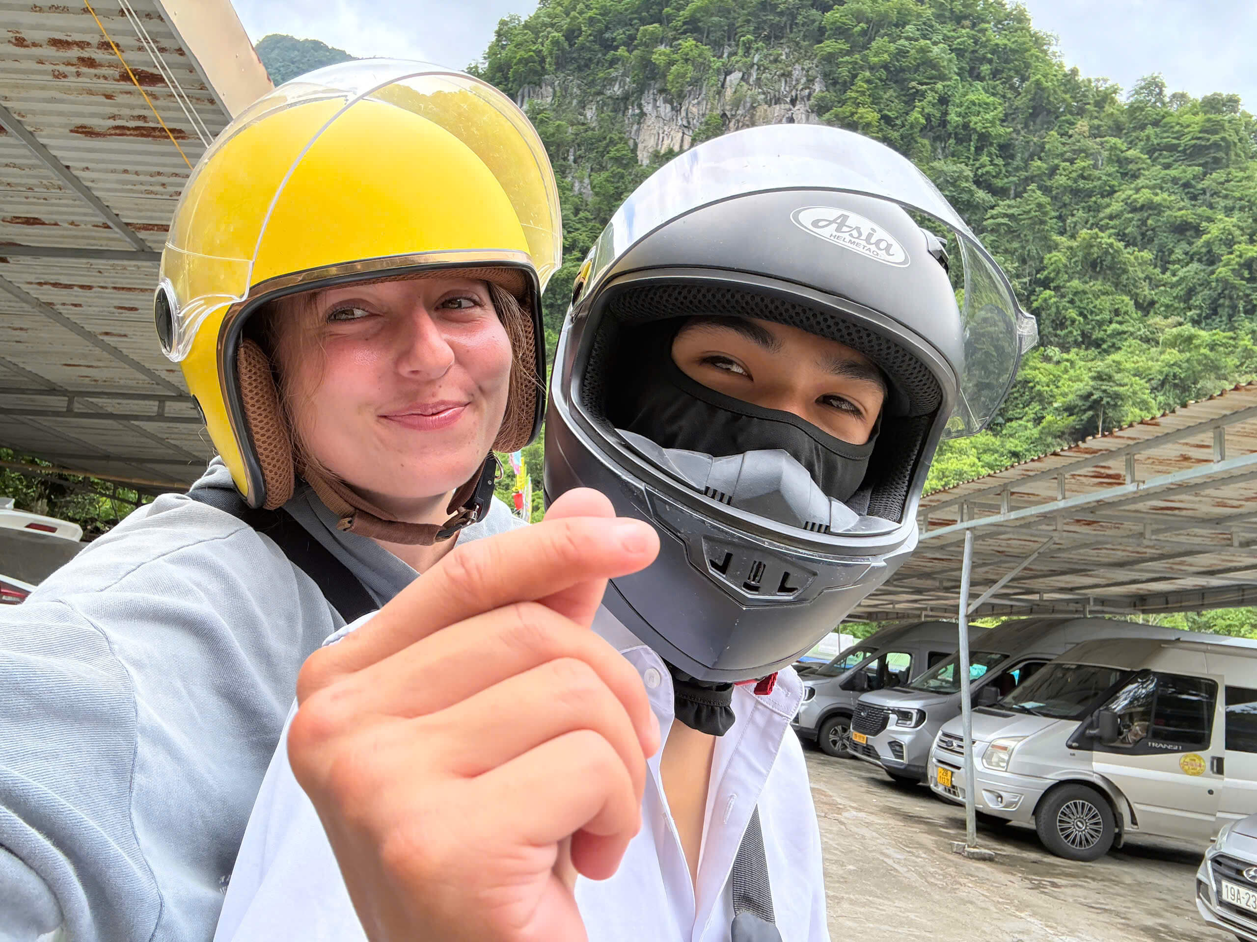 A smiling female tourist in a yellow helmet making a finger heart gesture while taking a selfie with her local Easy Rider guide, who is wearing a protective black full-face helmet.